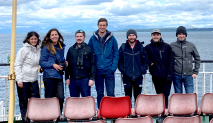7 students standing in row leaning against a rail with sea behind them
