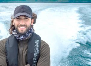 Luke Evans portrait photo wearing coat and baseball cap, life jacket on a boat with waves and sea in the background